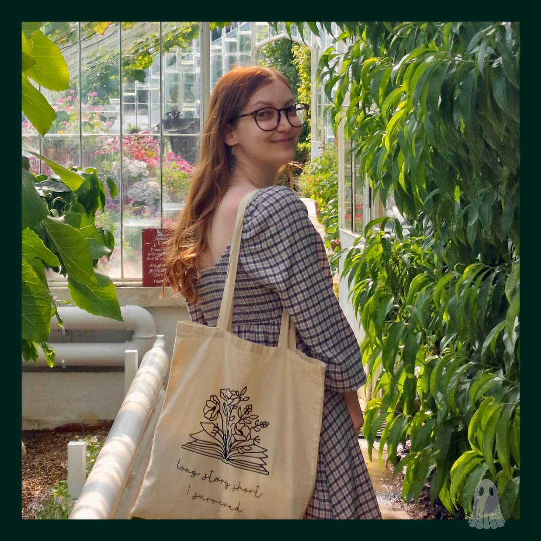 Picture shows the natural cotton canvas tote bag with 'long story short i survived' design, open book with flowers, modelled by Keeley, a white girl with long wavy auburn hair, glasses, and a brown checkered print dress in a botanical garden
