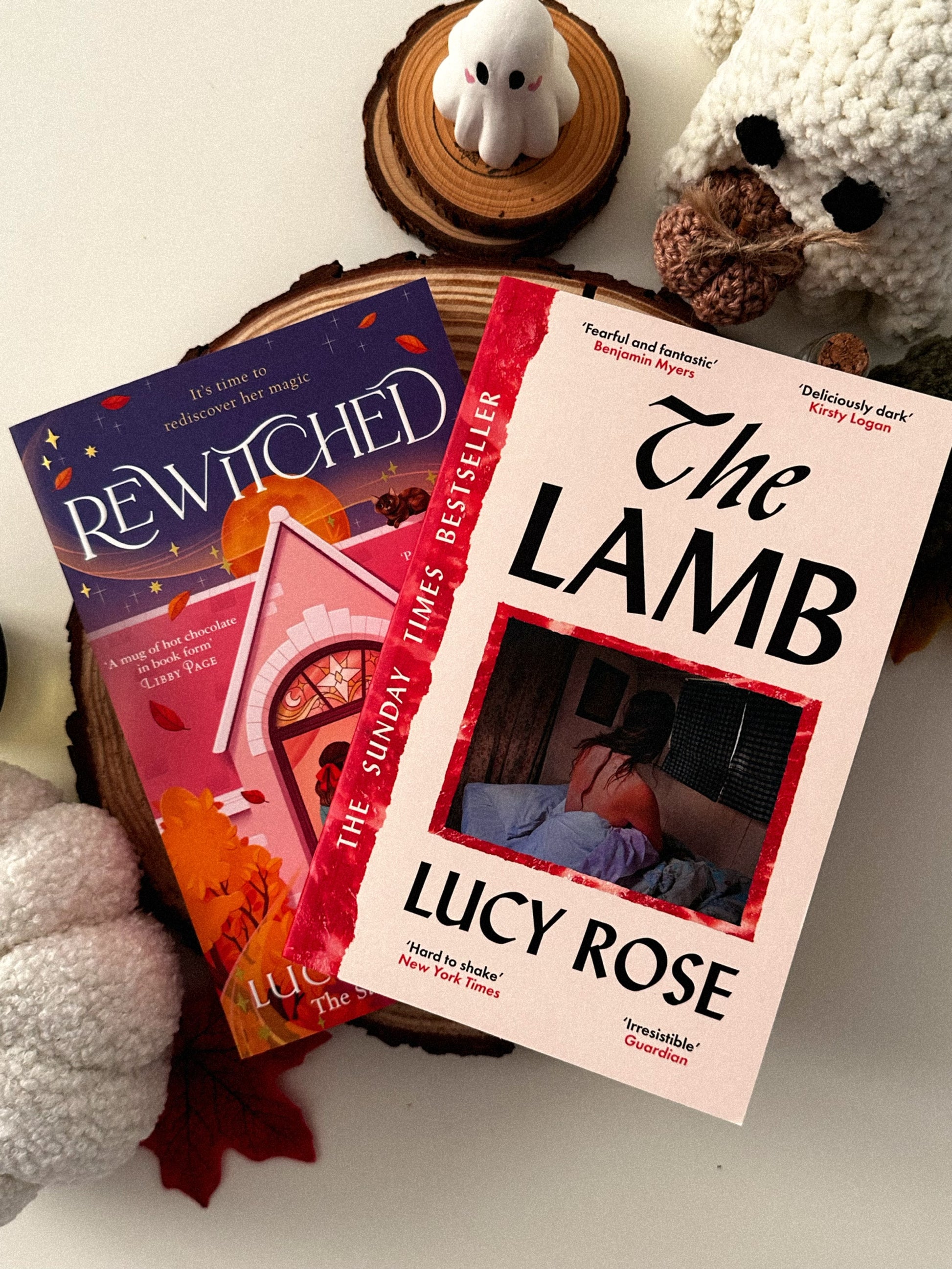 A flatlay of two books (The Lamb by Lucy Rose and Rewitched by Lucy Jane Wood) on a wood slice, with some decorative Halloween items - including a clay ghost, a crochet ghost, and a boucle pumpkin.