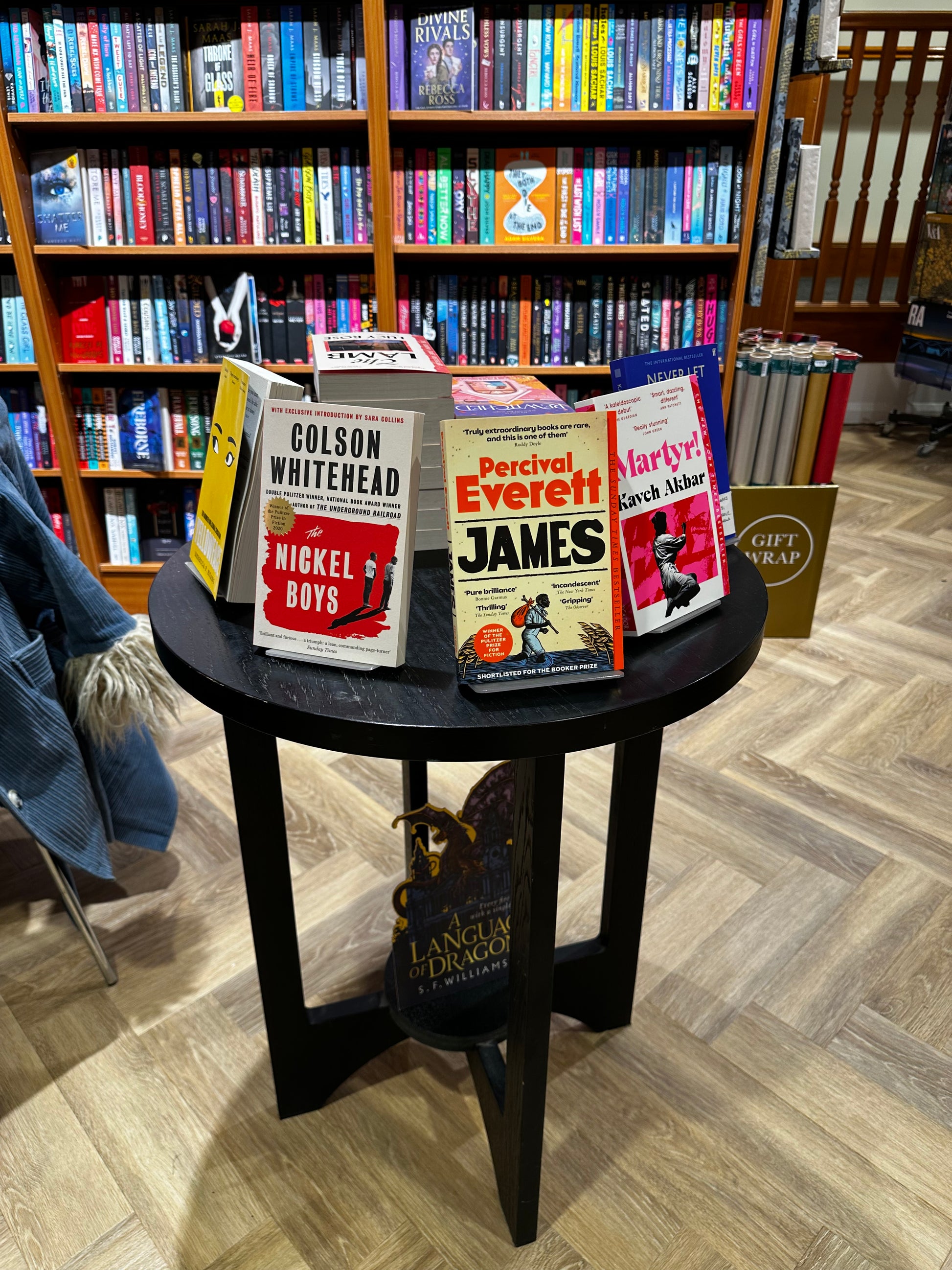 A selection of books on a wooden table in front of a bookshelf with a herringbone pattern floor. This is the display of books for our November Book Club Vote.